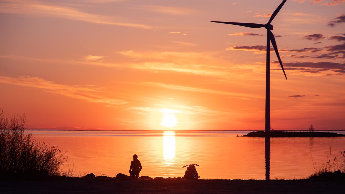 Ihminen seisoo rannalla auringonlaskussa. Kuvan oikeassa reunassa näkyy tuulivoimala.