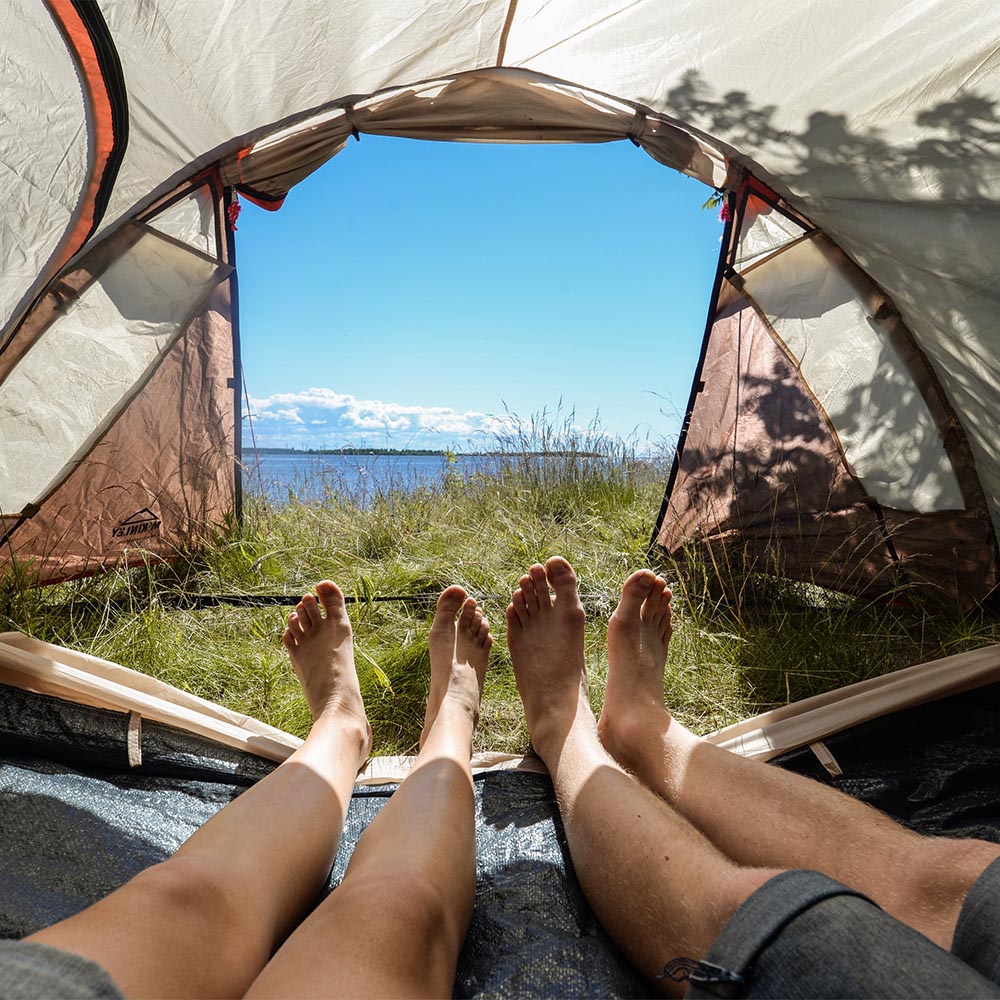 Aurinkoiselle rannalle pystytetty teltta, jonka sisäpuolelta otetussa kuvassa näkyvät kahden teltassa makaavan ihmisen jalat